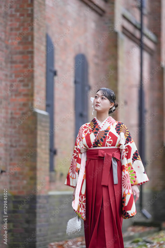 A Japanese woman in her 20s, wearing a red kimono (hakama), a staple of Japanese university graduates, walks down a path filled with autumn leaves in front of a traditional Japanese brick building.