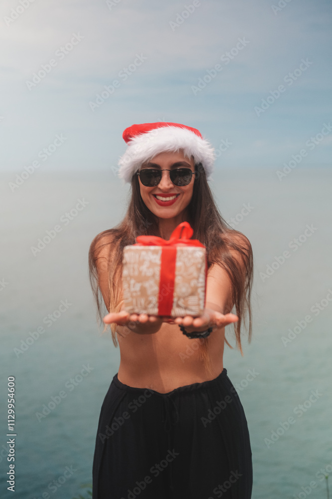 Obraz premium Portrait of young brunette woman in sunglasses, red bikini and Santa hat holding gift box with bow on ocean background. Tropical Christmas and New Year in summer on island. Holiday surprise.