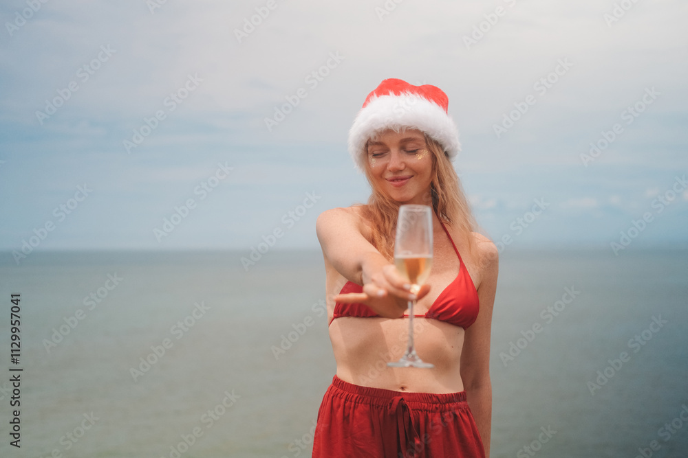 Obraz premium Banner with young red haired woman in Santa hat, red bikini holding glass of champagne standing on the coast against ocean background. Celebrating Christmas and New Year in summer on tropical island.