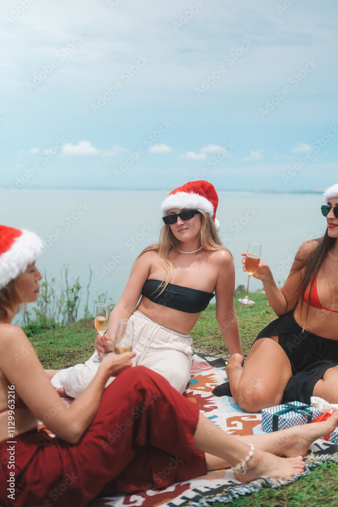 Obraz premium Young women friends brunette and blonde sitting on Christmas picnic in Santa hats and sunglasses on the ocean background and holding a glass of champagne in their hands. Tropical New Year on vacation.