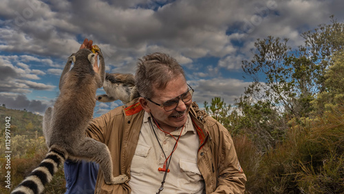 Ring-tailed lemurs catta have climbed onto a man's shoulder, reaching for a banana in his hand. The man laughs. The background is green vegetation, blue sky, clouds. Madagascar.   Nosy Soa Park photo