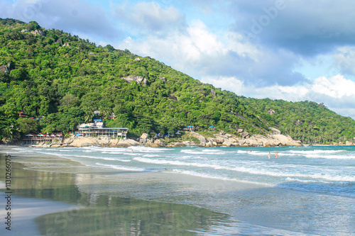 Famous Haad Rin Beach in Koh Phangan with white sandy shoreline, rolling waves, and beachfront resorts nestled against green hillside with granite boulders photo