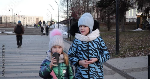 Young sisters dressed in warm winter clothing walking down steps in urban outdoor setting. Tracking camera movement following their descent, wide-angle composition to capture steps, surroundings, city