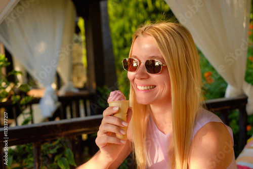 Blonde girl with bright smile and sunglasses on vacation eating pink ice cream in a cone. Joyful woman enjoying ice cream in park, copy space.