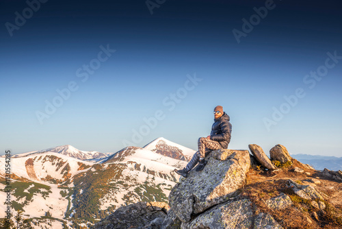 Stylish man with a beard and curled mustache enjoying view from top of a rocky mountain at sunrise against a scenic backdrop of Hoverla peak