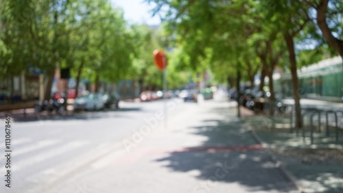 Blurred street with trees, cars, and a defocused traffic sign on a sunny day capturing a typical urban scene with green foliage and indistinct sidewalk environment.