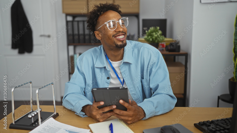 Fototapeta premium Handsome african american man with a beard smiling in an office environment holding a tablet, surrounded by paperwork and office supplies.