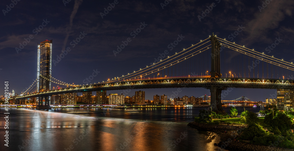 Fototapeta premium The photo captures the Manhattan Bridge at night from Brooklyn, with illuminated city lights reflecting on the calm water. The skyline, a modern skyscraper, and the serene park enhance the scene.