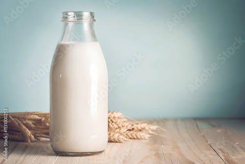 A glass milk bottle stands on a rustic wooden table with wheat stalks in the background and a soft blue backdrop. The composition offers ample copy space, highlighting its simplicity. photo