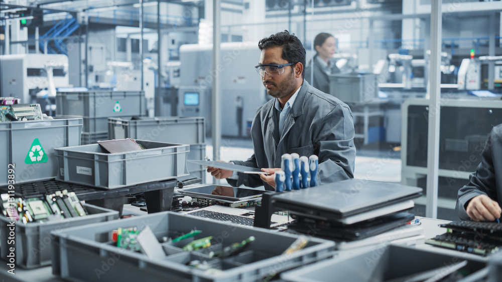 Fototapeta premium Indian Male Worker Disassembling Old Laptops To Recycle Electronic Components For Microchip Production At an Automated Electronics Factory With Robotic Arms. Man Unscrewing Displays And Sorting Them.