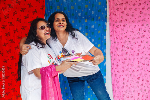 Two joyful Indian women stand playfully, holding a plate filled with vibrant colored powder, celebrating the lively Indian festival of colors, Holi. photo