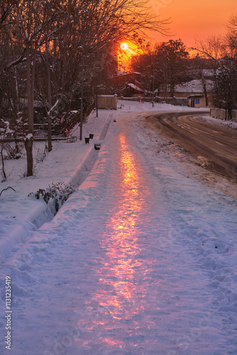 Urban sunset landscape with a snow-covered sidewalk where the setting sun is reflected, creating a beautiful light path on the ice