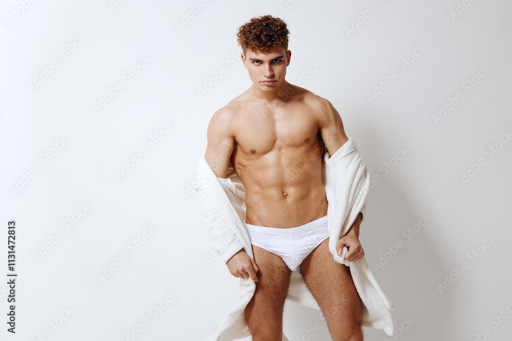 Fototapeta premium Confident young man posing in minimalistic studio setting, wearing white underwear, showcasing fitness and body positivity. Body empowerment and self-expression concept.