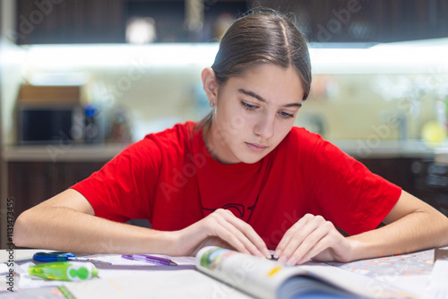 Young girl studying with focus at a table. A young girl wearing a red shirt studies intently at a table, surrounded by stationery items.