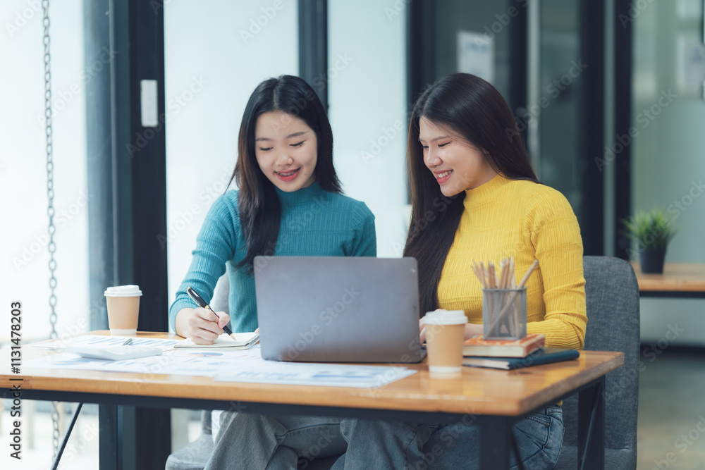 Obraz premium Collaborative Work: Two young women, dressed in casual attire, work together on a project, demonstrating focus and teamwork. They are engaged in a discussion, looking at a laptop, and taking notes.