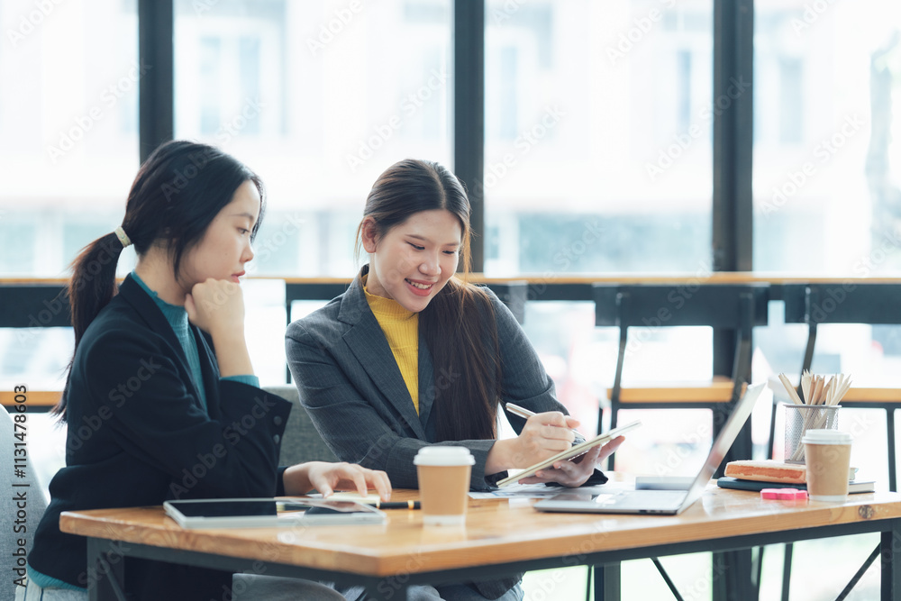 Strategic Collaboration: Two young Asian businesswomen engage in a productive discussion, reviewing documents and utilizing laptops in a modern, bright office setting.  