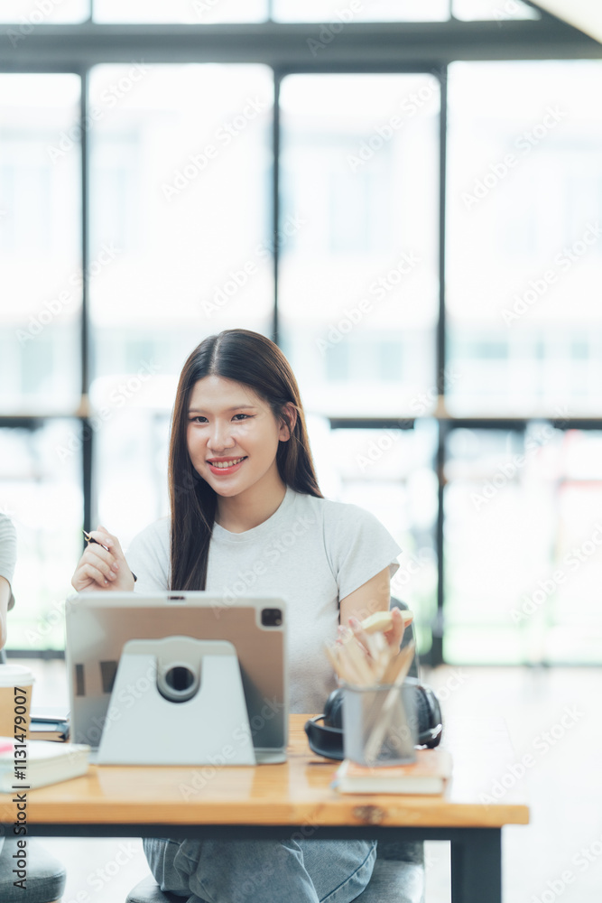 Obraz premium Focused and Inspired: A young woman, radiating positivity, works on a tablet in a modern office setting, showcasing the power of technology and creativity. 