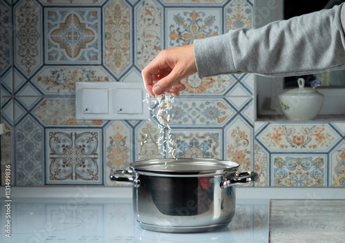 A woman's hand throws grains of rice into a pot of boiling water on the stove. Cooking food.