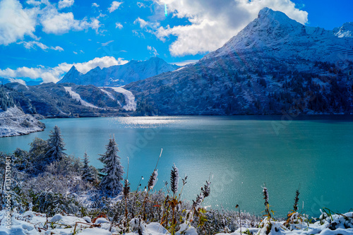 A view of the picturesque alpine lake the Kops Reservoir with green water and reflections, surrounded by snow covered mountains. In the foreground - snow-covered trees and plants photo