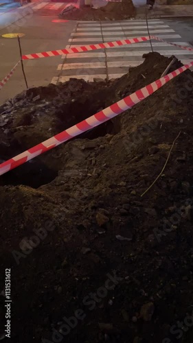 Night excavation work is ongoing, with warning tape marking the nearby pedestrian area for safety. Vertical photo