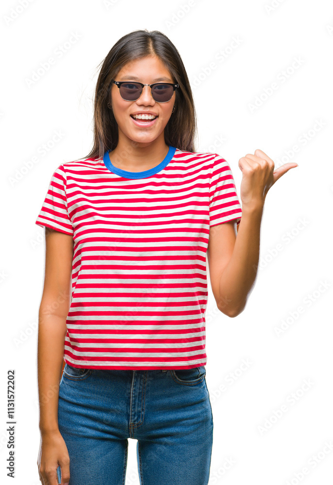 Young asian woman wearing sunglasses over isolated background smiling with happy face looking and pointing to the side with thumb up. Young asian woman wearing sunglasses over isolated background smiling with happy face looking and pointing to the side with thumb up.