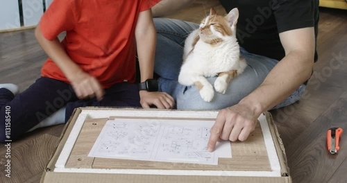Dad and son sit looking at instructions for assembling nightstand. Cat lies on father crossed legs. Man and young boy assemble furniture
