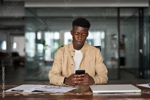 Busy professional African business man sitting in office holding mobile cellphone. Young Black businessman entrepreneur using smartphone looking at cell phone technology device at work.