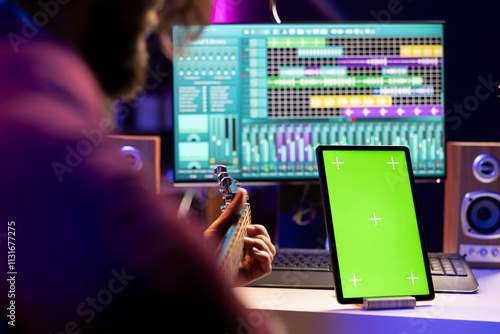 Music producer guitarist playing acoustic instrument in home studio, recording a new song on daw software interface. Artist learns new guitar accords next to greenscreen display tablet. photo