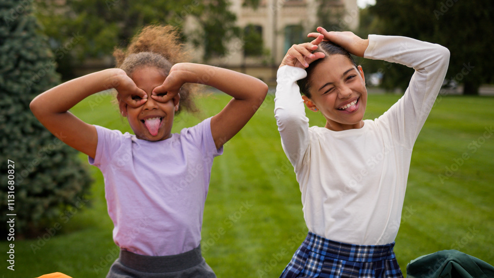 Obraz premium Happy little girls two kids schoolgirls children school education students pupils classmates multiracial African Indian female friends laugh have fun eyes circles grimace play game kidding outdoors