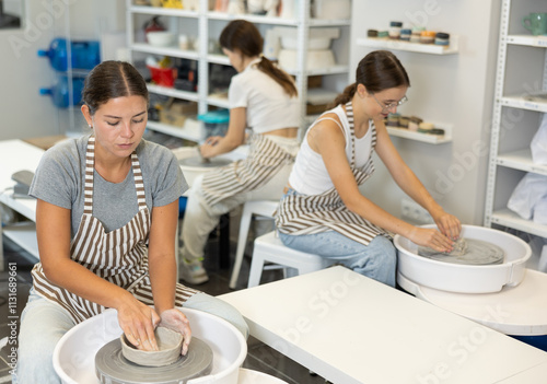 Three young women sits near potter wheel and makes clay crafts, makes vase plate, works in company of like-minded craftsmen. Handmade art, hobbies and creativity. photo