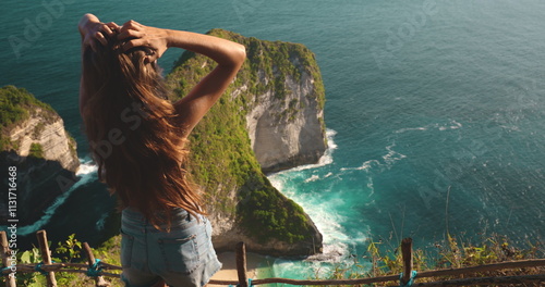 A woman stands on top of a cliff overlooking the vast ocean on Nusa Penida Island in Indonesia. The scene captures her in contemplation as she takes in the breathtaking view below. photo