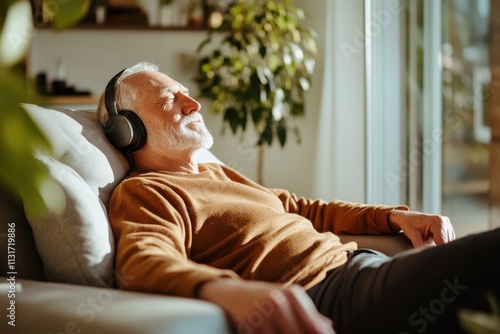 An elderly man in a brown sweater is lounging on a couch, headphones on, bathed in sunlight while enjoying his favorite tunes in a bright, modern room. photo