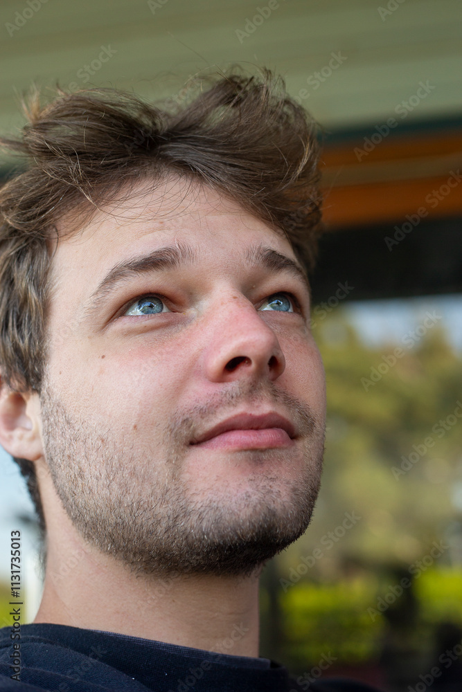 Fototapeta premium A close-up of a young man's face with vibrant blue eyes looking upward. The photo captures fine skin details, soft natural light, and a dreamy, introspective mood with a blurred green background