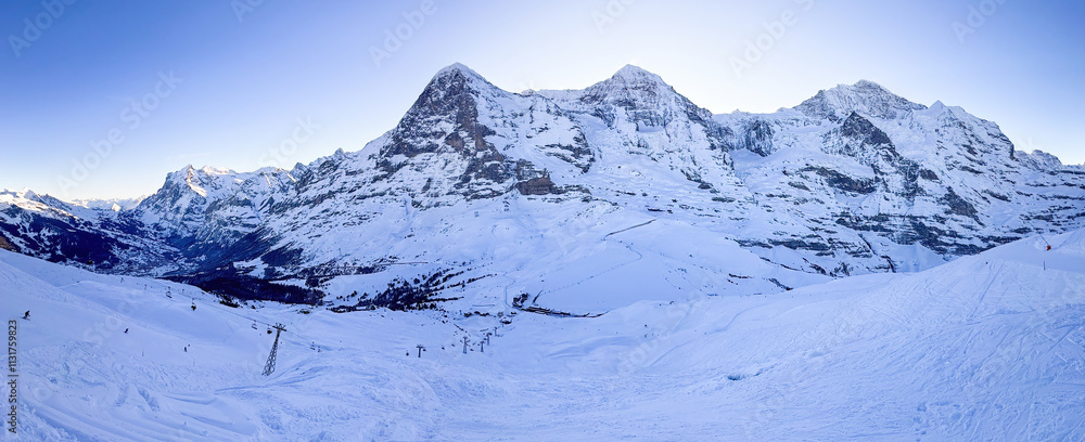 Panoramic view of famous snowcoverd mountains Eiger, Moench and Jungfrau (from left to right) in Switzerland with Grindelwald and Wetterhorn (far left) in winter early in the morning against blue sky