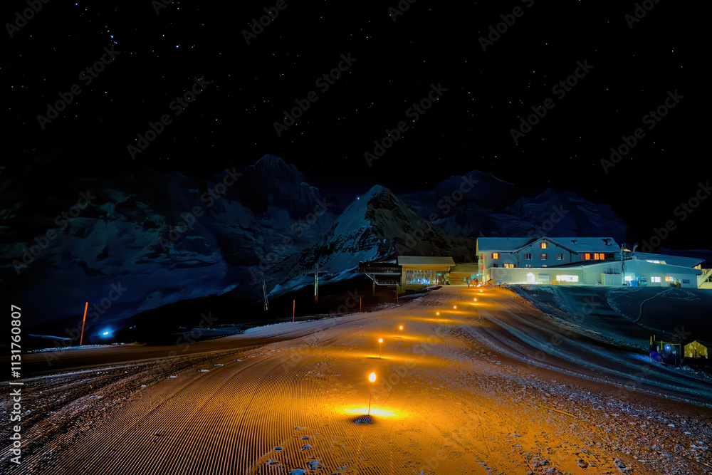 Torches showing the way to Berghaus Maennlichen at night in winter, Grindelwald, Switzerland with Moench, Tschuggen and Jungfrau mountain in the background against sky full of stars