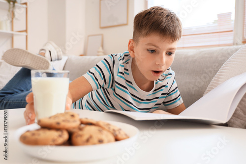 Shocked little boy with glass of milk reading magazine on sofa at home