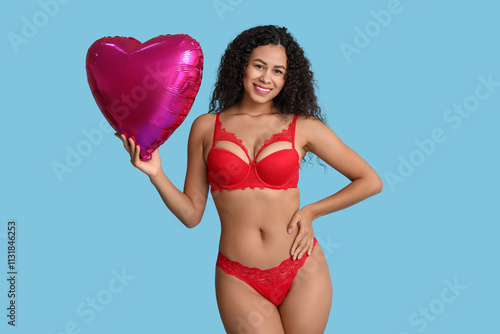 Beautiful young African-American woman in stylish red lingerie with air balloon in shape of heart on blue background. Valentine's Day celebration