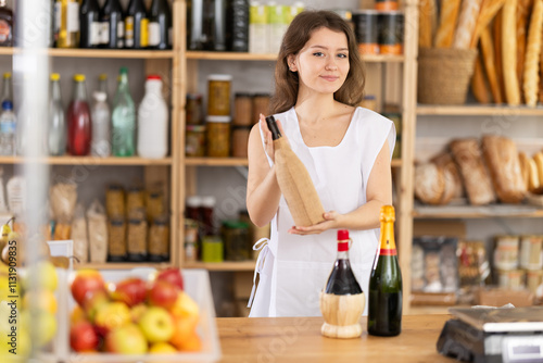 Positive young saleswoman is holding a bottle of wine standing at the counter in market photo