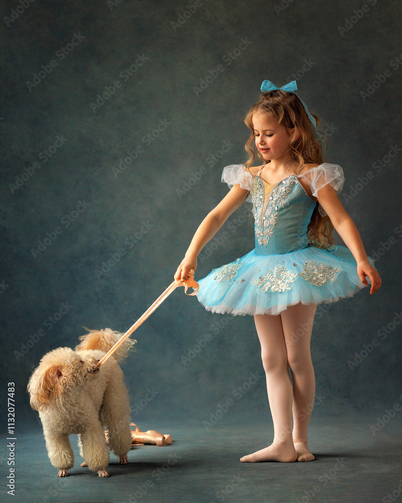 A ballerina girl in a ballet skirt and pointe shoes poses with poodle dog. Little adorable child ballerina in ballet tutu with puppy practicing classic choreography in ballet school, in studio