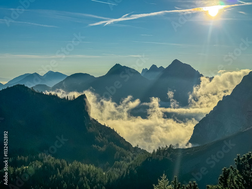 Sun rays pierce through clouds casting warm glow on mountain peaks on Loibl Pass in Karawanks, Slovenia Austria. Dramatic interplay of light and shadow. Jagged edges against hues of morning dawn. Hike photo