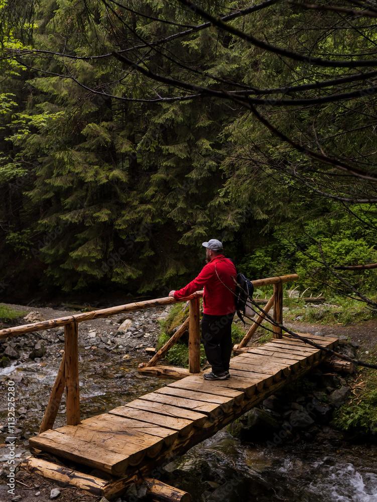 Fototapeta premium Man sitting on a bridge in the forest with a rocky path along a river. Man taking a break at the bridge by the river. Active vacation concept.