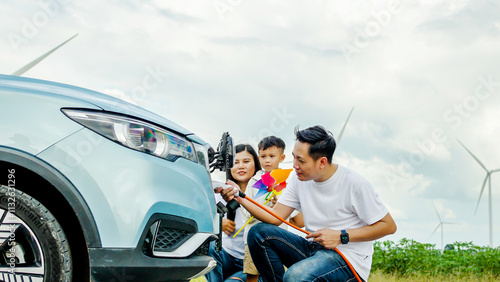 Asian family charging the ev car with eco-friendly, windmill in background, Electric vehicle driven by clean renewable energy from wind turbine generator to charger station. photo