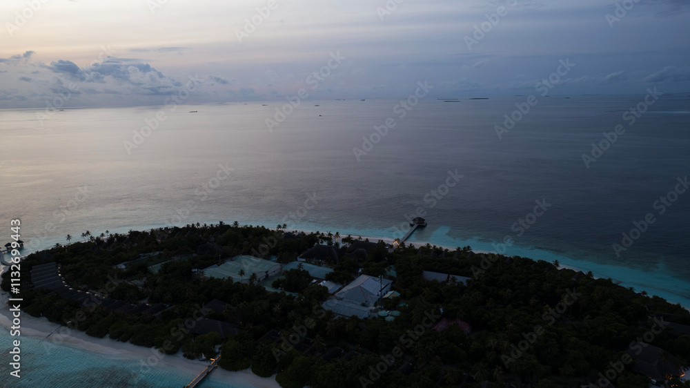 Top view of the sunset over an island in the ocean. Large dark clouds with a orange tinge. Turquoise ocean water, white sand of the beach. Green palm trees. Villas on the water. Water is like a mirror