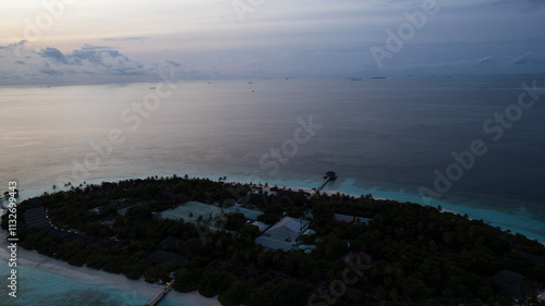 Obraz na płótnie Top view of the sunset over an island in the ocean. Large dark clouds with a orange tinge. Turquoise ocean water, white sand of the beach. Green palm trees. Villas on the water. Water is like a mirror w salonie