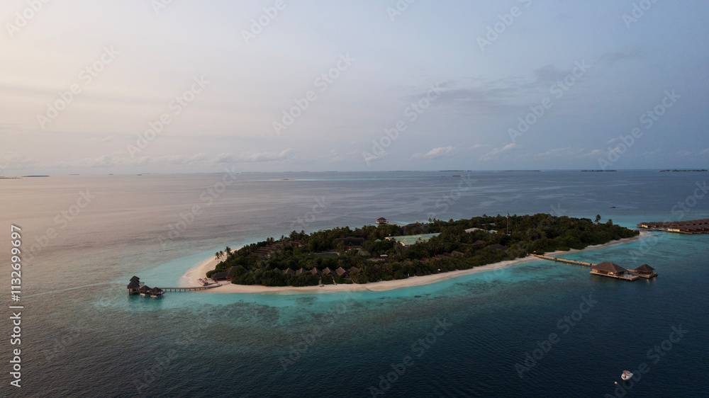 Top view of the sunset over an island in the ocean. Large dark clouds with a orange tinge. Turquoise ocean water, white sand of the beach. Green palm trees. Villas on the water. Water is like a mirror
