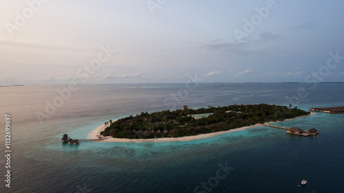 Top view of the sunset over an island in the ocean. Large dark clouds with a orange tinge. Turquoise ocean water, white sand of the beach. Green palm trees. Villas on the water. Water is like a mirror photo