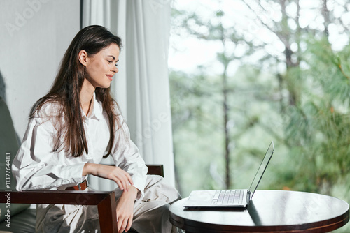 Young woman in a bright room working on a laptop, dressed smartly in white, showing a focused expression, with greenery visible through the window photo