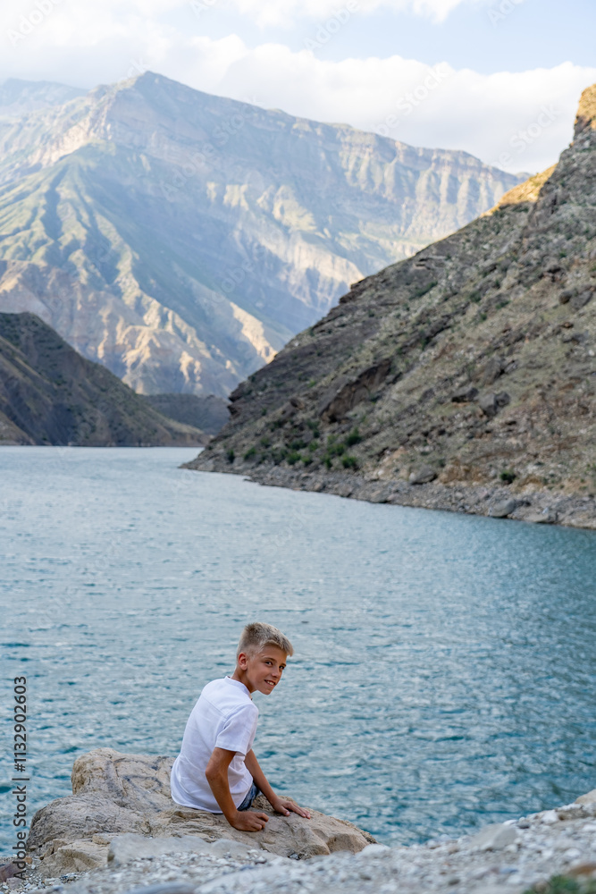 Fototapeta premium A young boy sits on a rocky shore, gazing at the tranquil lake surrounded by towering mountains in the warm light of late afternoon