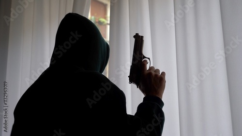 Vászonkép hooded terrorist prepares to use a revolver gun and shoot from the window toward