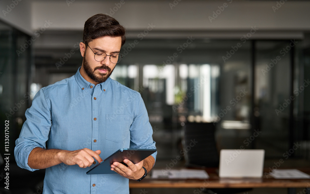 © insta_photos - Busy young Latin business man manager using tablet computer, serious hispanic businessman executive looking at tab device analyzing finance trading market data working standing in office.